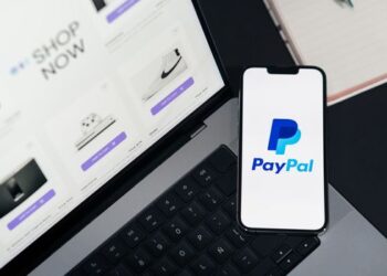 Smartphone displaying PayPal logo next to laptop with online shopping site open.