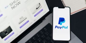 Smartphone displaying PayPal logo next to laptop with online shopping site open.
