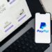 Smartphone displaying PayPal logo next to laptop with online shopping site open.